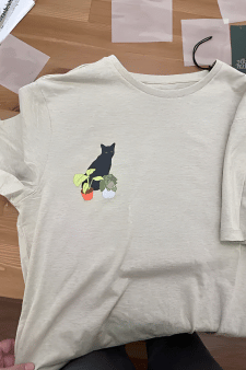 Minimalistischer DTF Transfer auf einem beigen T-Shirt: Eine kleine schwarze Katze sitzt zwischen zwei Zimmerpflanzen – ein dezentes und hochwertiges Bügelbild für Katzenliebhaber.